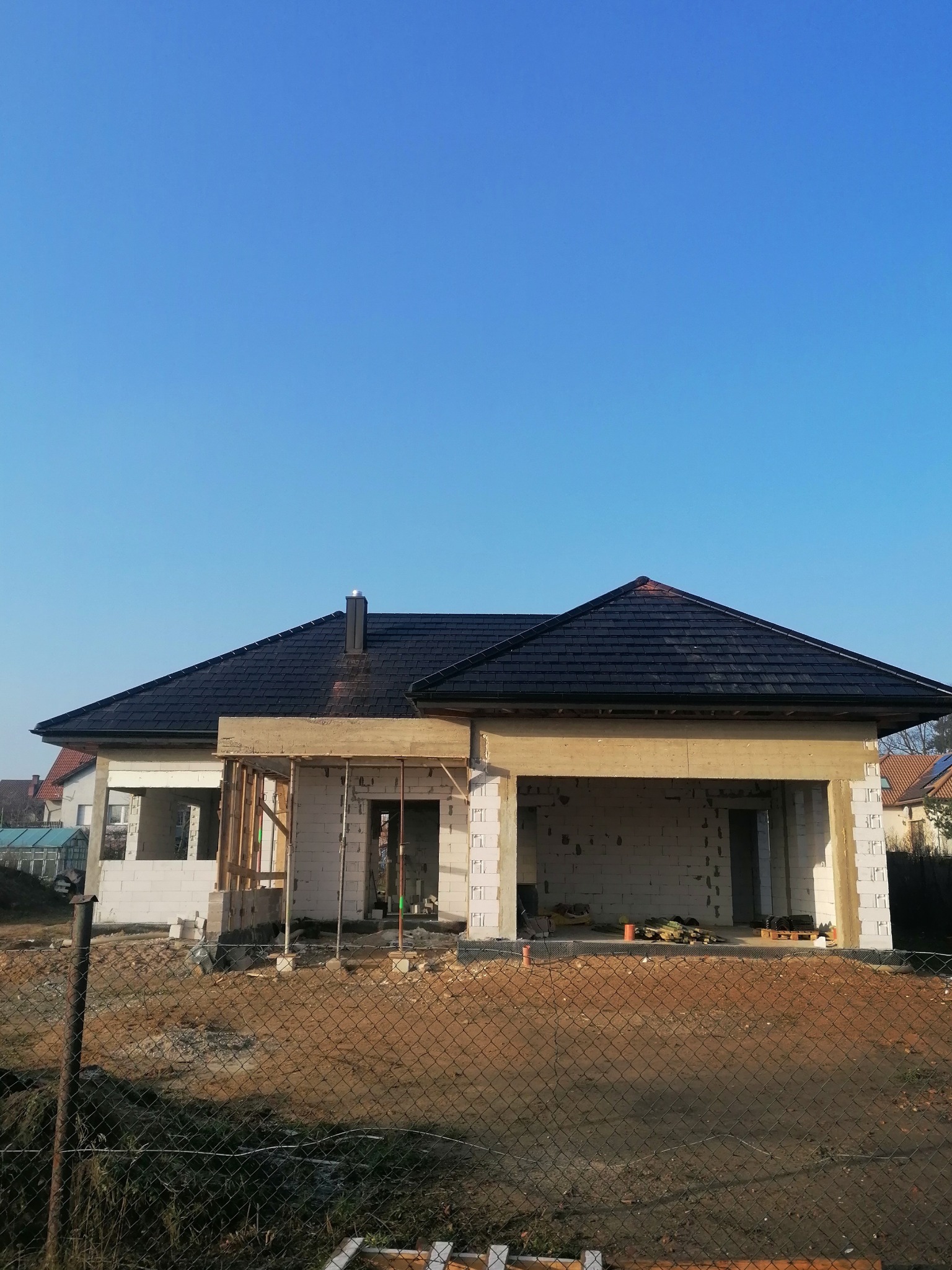 Dom w trakcie budowy z czarnym dachem z dachówki ceramicznej, widok zza ogrodzenia, konstrukcja w stanie surowym otwartym.