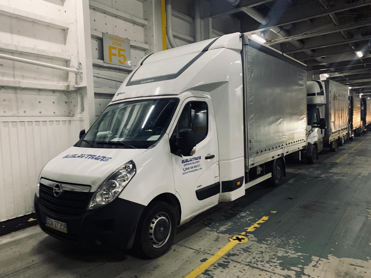 Kilkanaście białych busów transportowych z plandekami, ustawionych jeden za drugim w rzędzie na promie, widoczny znak strefy parkowania F5