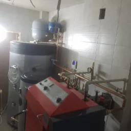 Zainstalowany kocioł zgazowujący drewno Atmos z buforem 800l i przygotowanymi podłączeniami do kotła gazowego, widoczne rury miedziane, manometry i sterownik na tle białych płytek.