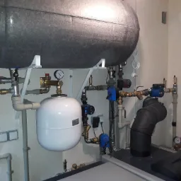 Złożona instalacja grzewcza z szarym zbiornikiem akumulacyjnym, białym naczyniem przeponowym, licznymi zaworami, rurami i pompami obiegowymi na tle jasnej ściany.