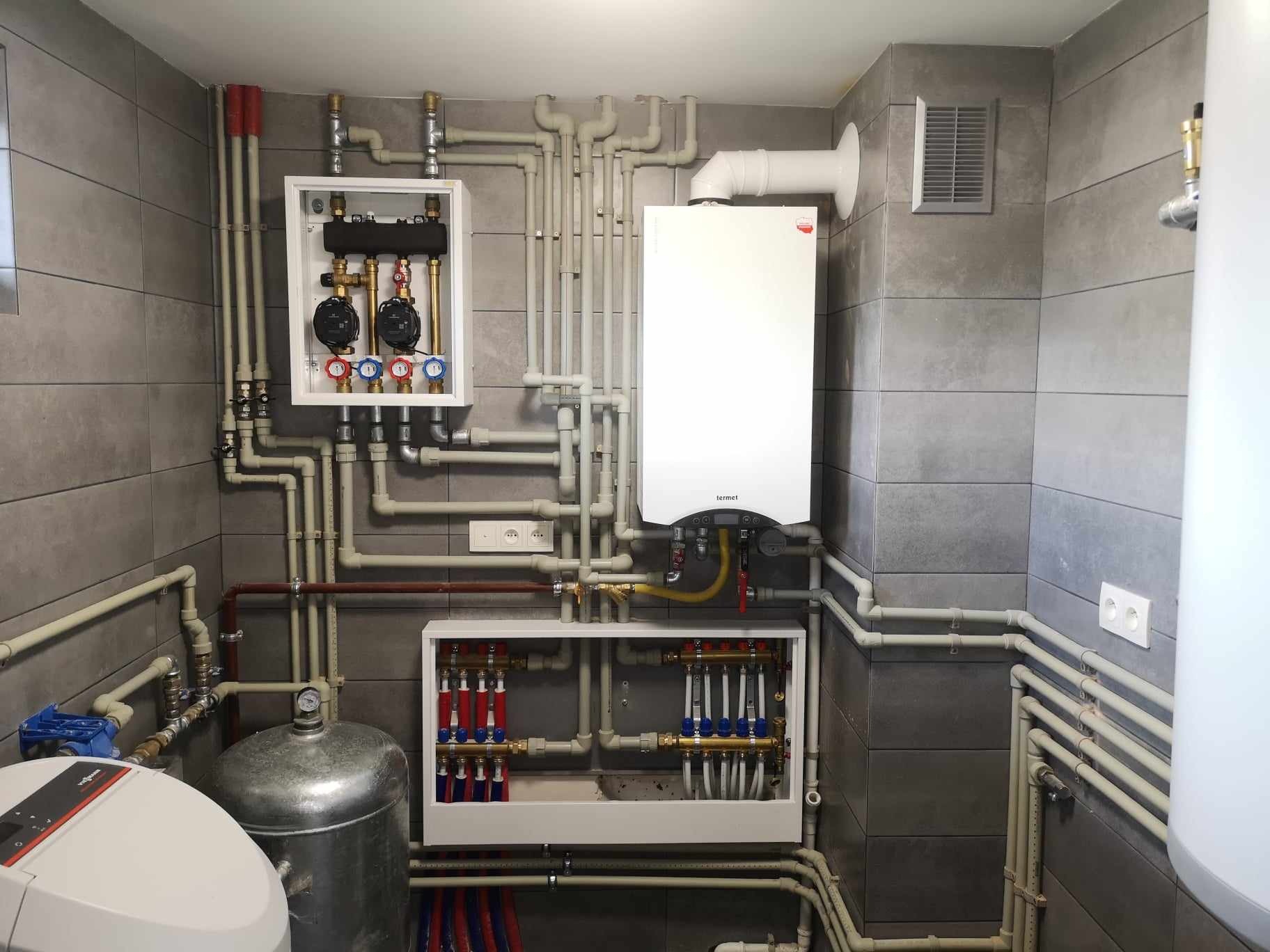 Węzeł cieplny z białym piecem gazowym Termet, rozdzielaczami, pompami obiegowymi i licznymi rurami na ścianie z szarych płytek.