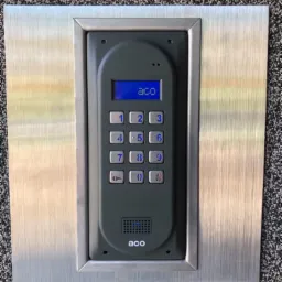 Szary panel domofonu ACO z metalową ramką, zamontowany na szorstkiej, ciemnej powierzchni.