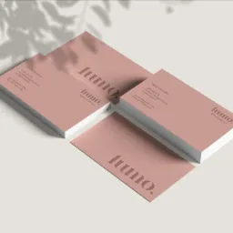 Stos wizytówek w odcieniach pudrowego różu z minimalistycznym logo 'luumo.' oraz danymi kontaktowymi, ułożony na jasnym tle z cieniem liści.