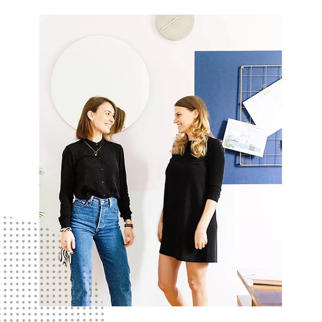 Dwie uśmiechnięte kobiety w biurze, jedna w dżinsach i czarnej koszuli, druga w czarnej sukience, rozmawiają na tle okrągłego lustra i tablicy z notatkami.