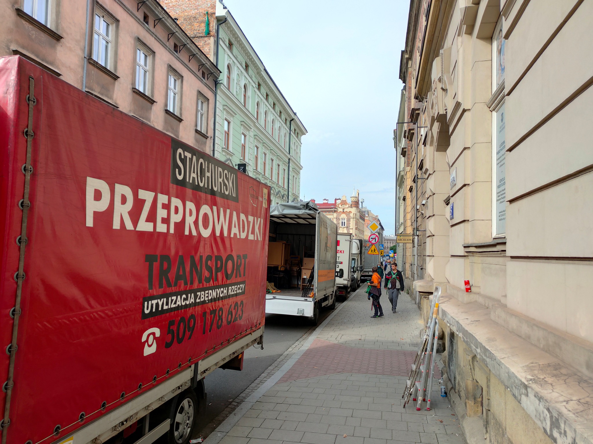 Czerwona plandeka samochodu ciężarowego z napisem 'STACHURSKI PRZEPROWADZKI TRANSPORT UTYLIZACJA ZBĘDNYCH RZECZY' i numerem telefonu na tle ulicy z budynkami w stylu kamienic oraz otwartych drzwi...