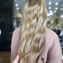 Długie, blond włosy z refleksami, widziane z tyłu, po zabiegu koloryzacji, na tle salonu z oświetleniem punktowym. W kadrze widoczna dłoń z czerwonym paznokciem.