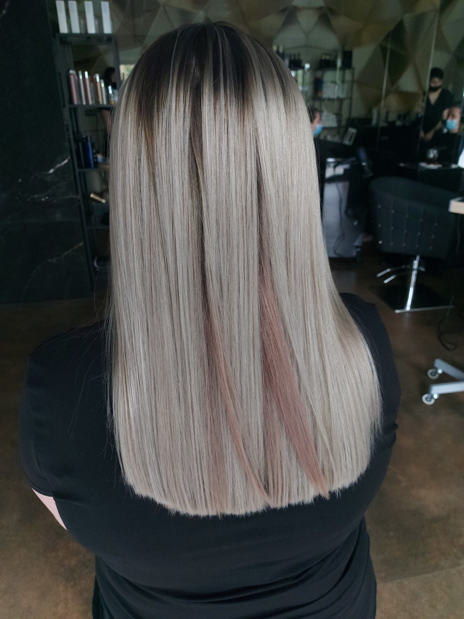 Proste, platynowe włosy z subtelnymi różowymi pasemkami, widoczne od tyłu, w tle salon z lustrami i stanowiskami pracy.