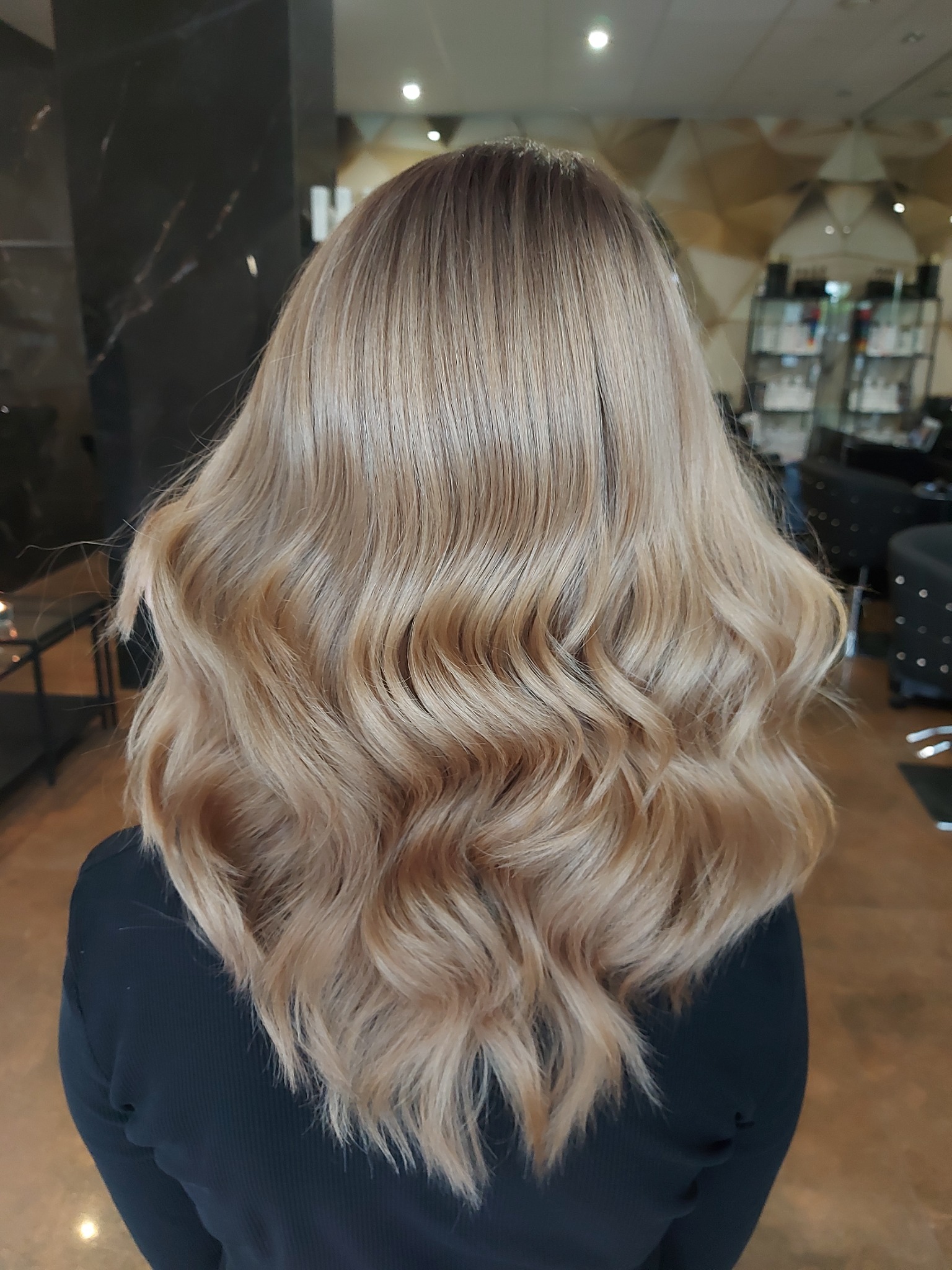 Tył głowy z długimi, blond włosami ułożonymi w delikatne fale, z widocznym gradientem koloru od ciemniejszego odrostu po jaśniejsze końcówki, na tle wnętrza salonu.