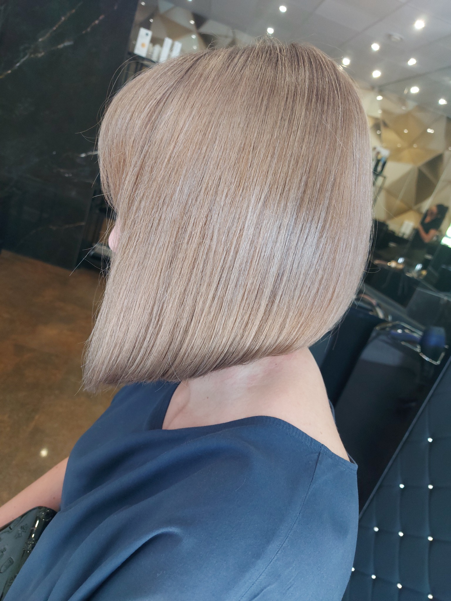 Blond bob z asymetrycznym cięciem, widoczne refleksy i gładka tekstura, zarys szyi kobiety w granatowej bluzce, tło salonu fryzjerskiego.
