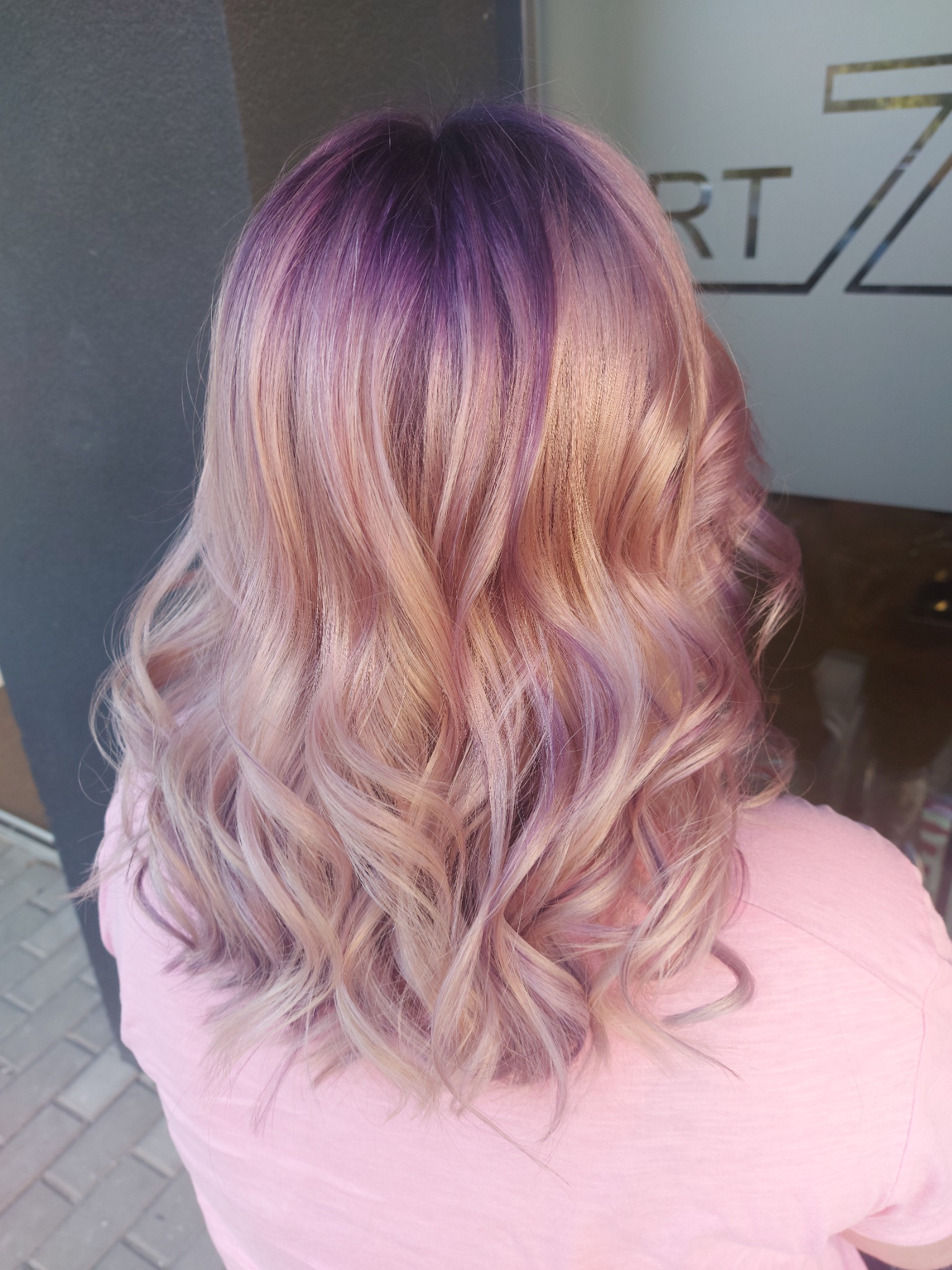 Włosy po koloryzacji z fioletowymi odrostami i różowo-blond refleksami, delikatnie pofalowane, widoczne od tyłu na tle ściany i szyby z logiem.