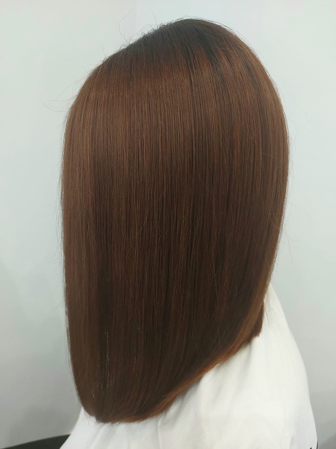 Brązowe, proste włosy do ramion, z ciemniejszym odrostem, widoczne od tyłu na tle białej bluzki i ściany.