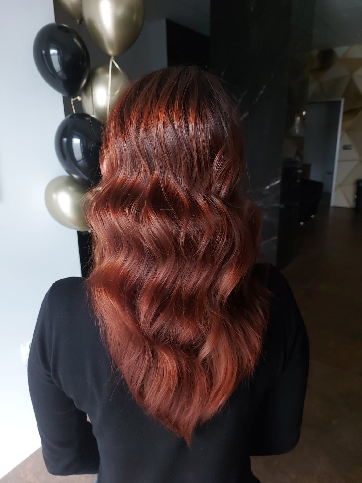 Widok z tyłu na falowane, miedziane włosy kobiety, z dekoracją z czarno-złotych balonów w tle, prawdopodobnie po koloryzacji i stylizacji.