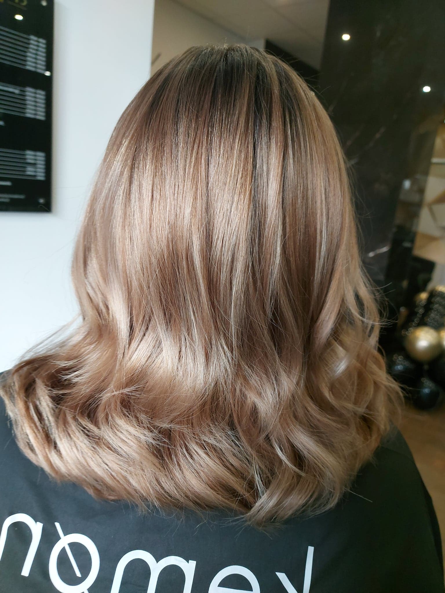 Widok z tyłu na falowane włosy w odcieniach blondu i brązu, z widocznym logotypem na czarnej koszulce osoby, stojącej na tle wnętrza salonu.