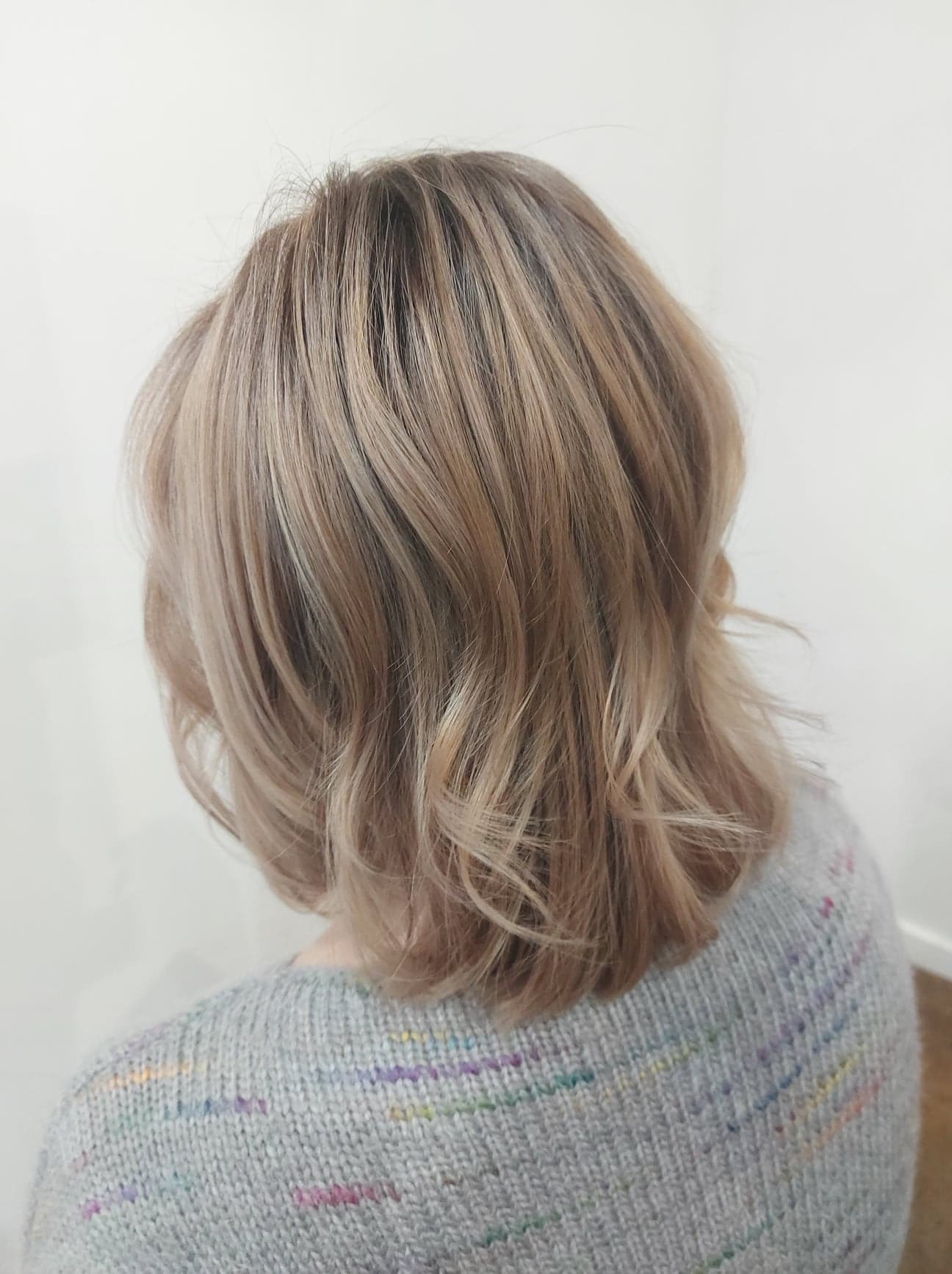 Krótkie, falowane włosy w odcieniach blondu z widocznymi refleksami, widziane od tyłu, na tle białej ściany i szarego swetra.