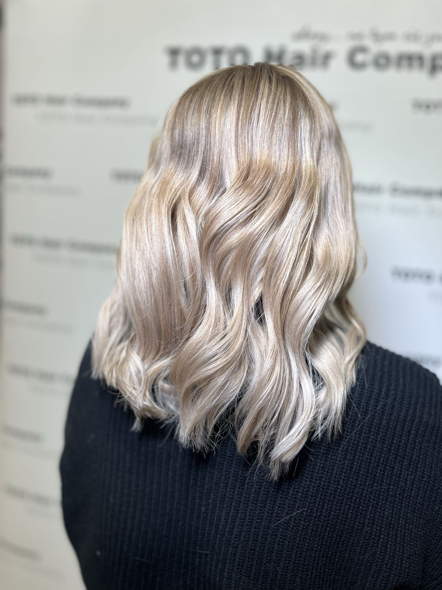 Tył głowy z platynowymi, pofalowanymi włosami, delikatne refleksy, czarny sweter, białe tło z logo firmy.