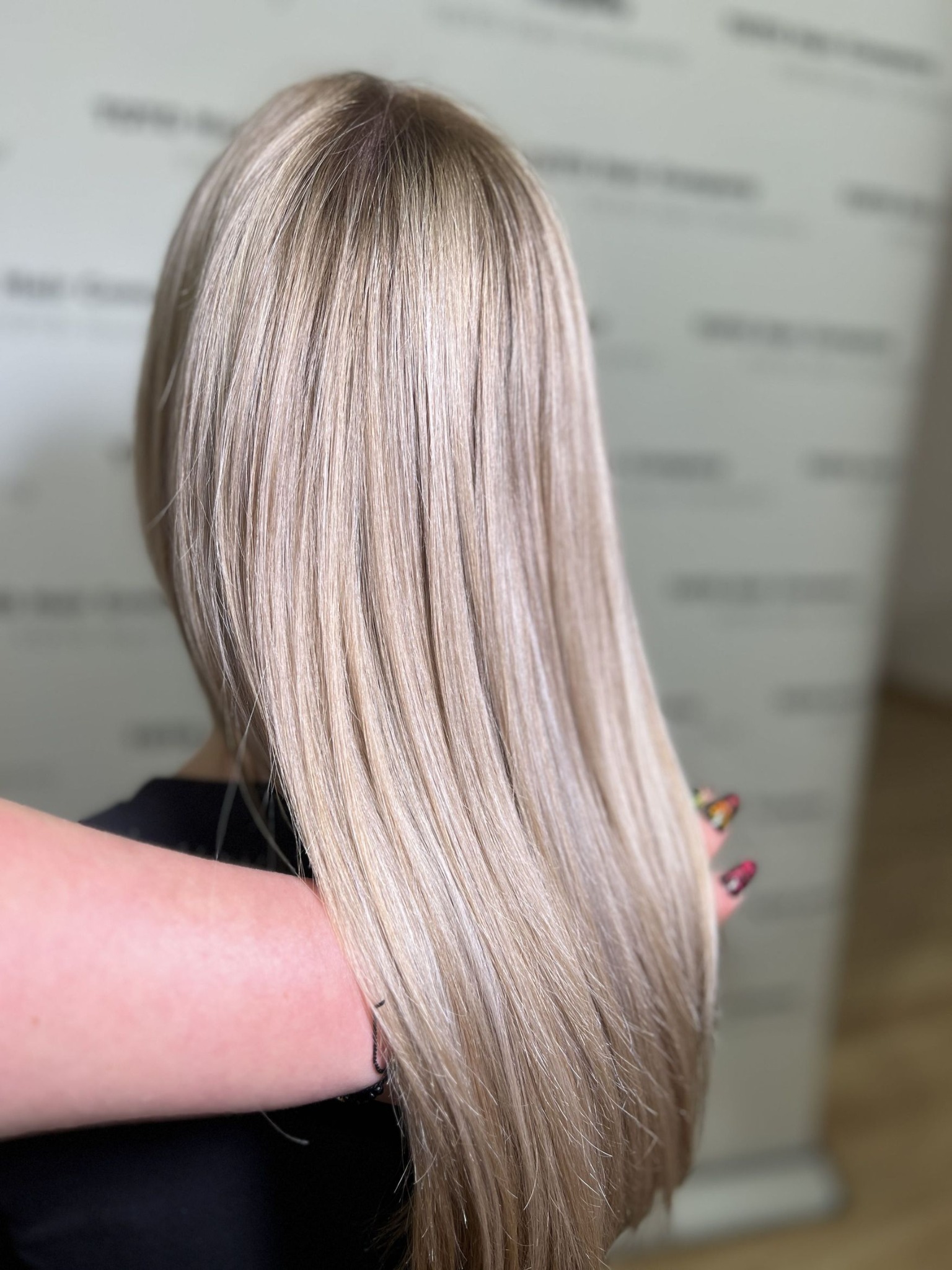 Długie, proste, blond włosy z delikatnym odrostem, prezentowane z tyłu, z dłonią w kadrze z kolorowym manicure.