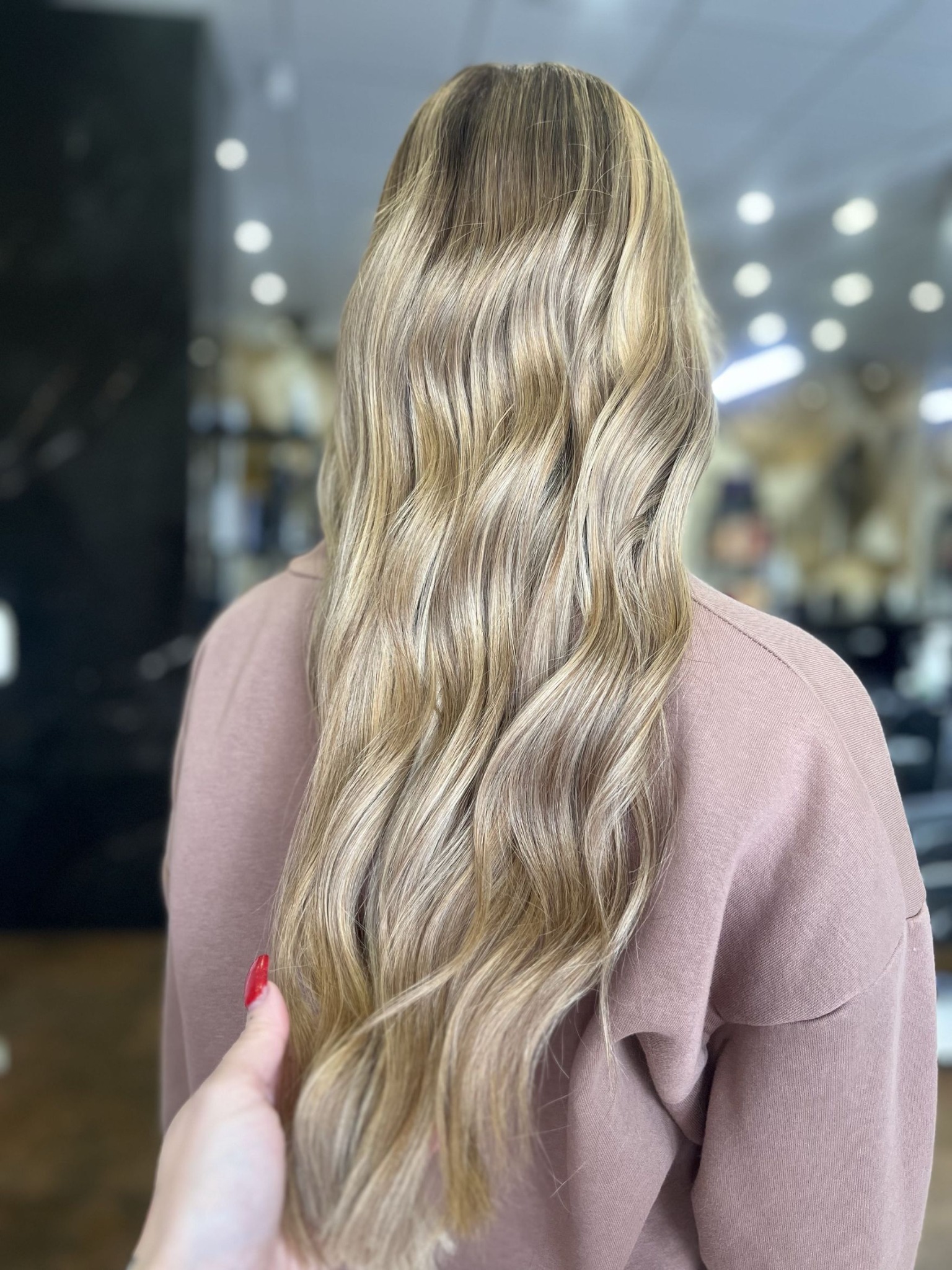 Długie, blond włosy z refleksami, widziane z tyłu, po zabiegu koloryzacji, na tle salonu z oświetleniem punktowym. W kadrze widoczna dłoń z czerwonym paznokciem.