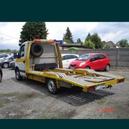 Adek-trans - Żółto-biała platforma pojazdu z zapasowym kołem i napisem 'POMOC DROGOWA' na dachu, zaparkowana na żwirowym parkingu z widocznymi samochodami osobowymi w tle.