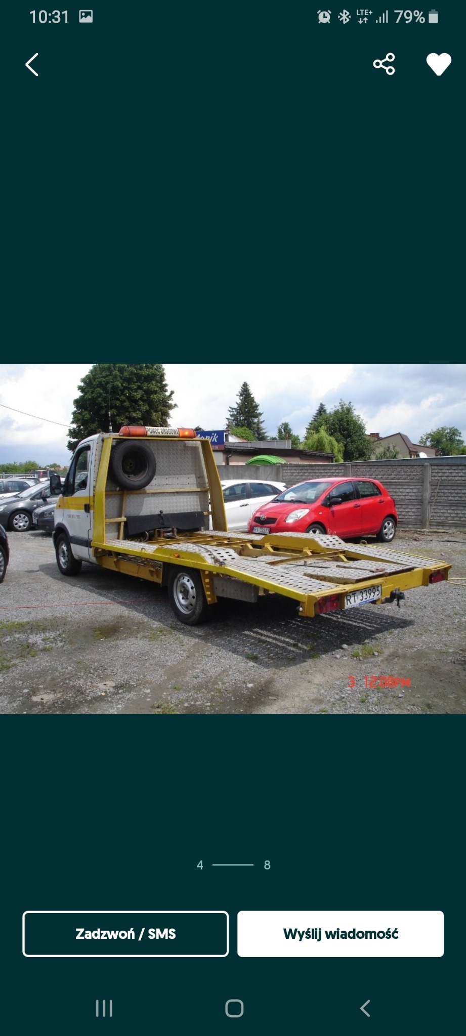 Żółto-biała platforma pojazdu z zapasowym kołem i napisem 'POMOC DROGOWA' na dachu, zaparkowana na żwirowym parkingu z widocznymi samochodami osobowymi w tle.