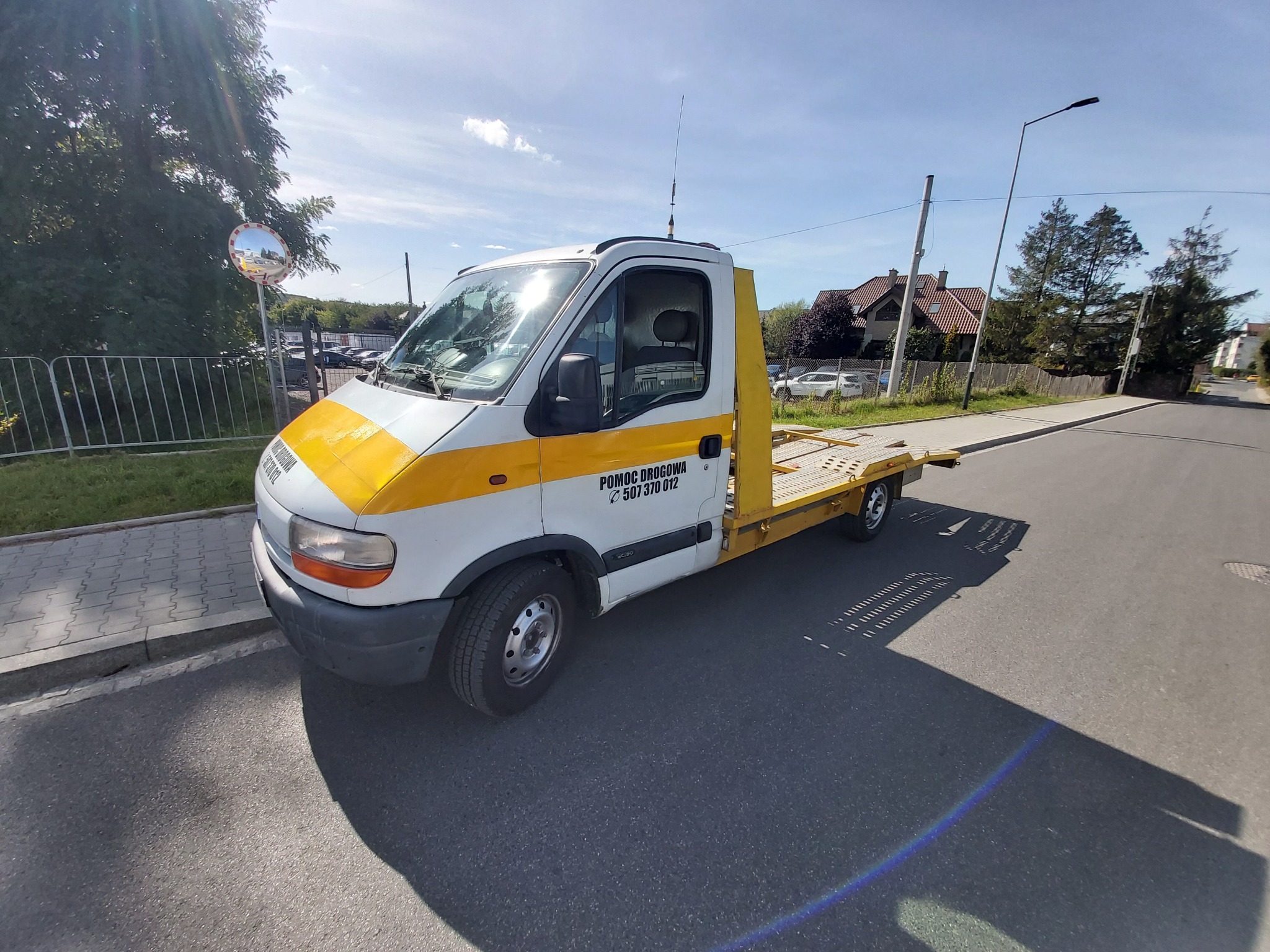 Żółto-biała laweta z napisem 'POMOC DROGOWA' zaparkowana na asfalcie w słoneczny dzień, widok z boku.