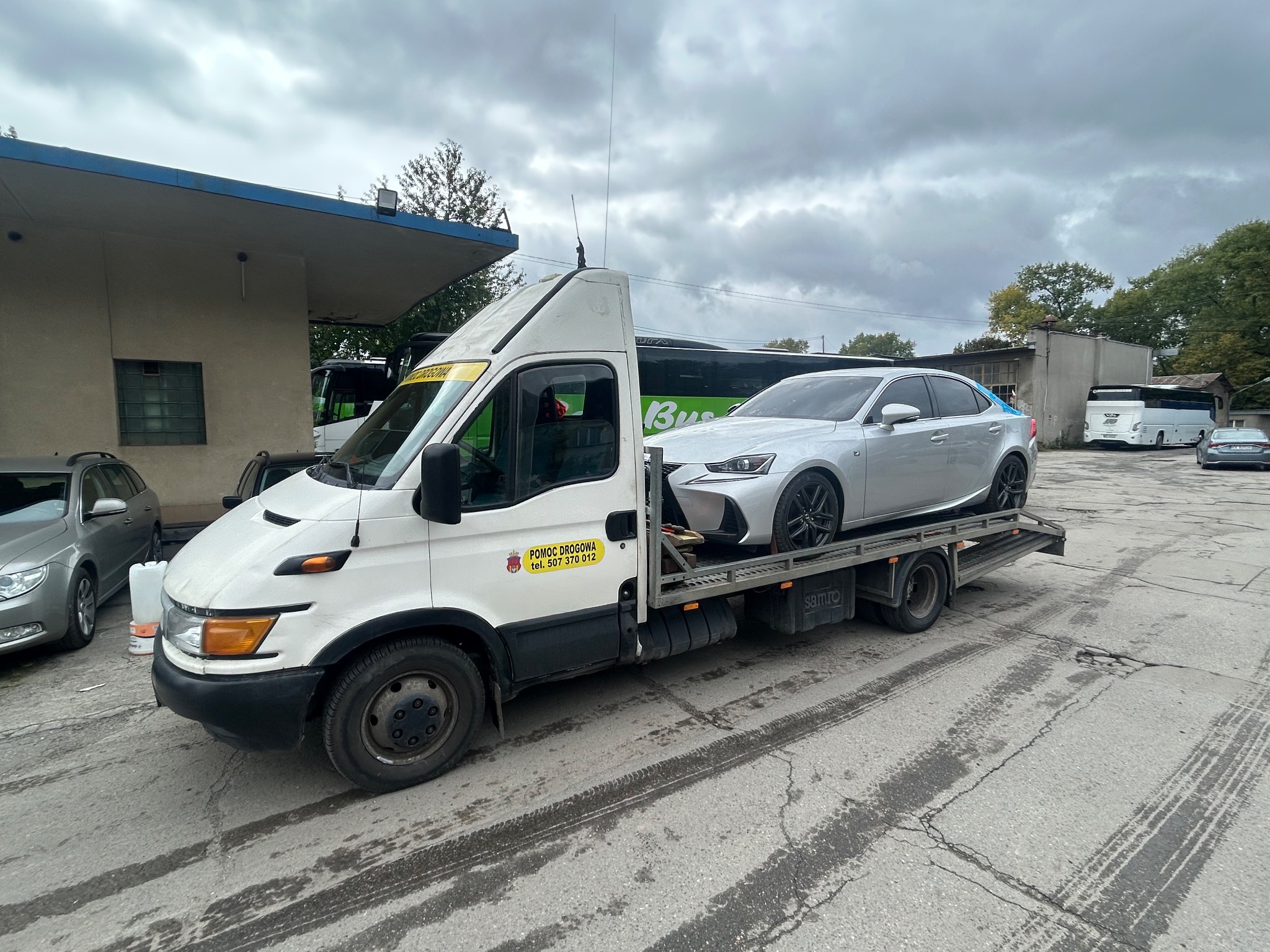 Biała laweta Iveco z srebrnym samochodem osobowym Lexus na platformie, zaparkowana na zewnątrz, w tle autobusy i pochmurne niebo.