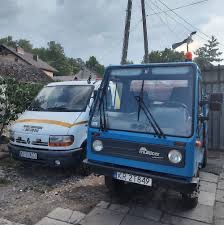 Dwa pojazdy: biały bus z żółtym pasem i niebieski pojazd specjalny z numerem rejestracyjnym KR 2T849 stojące obok siebie na brukowanym podjeździe, w tle domy i drzewa.