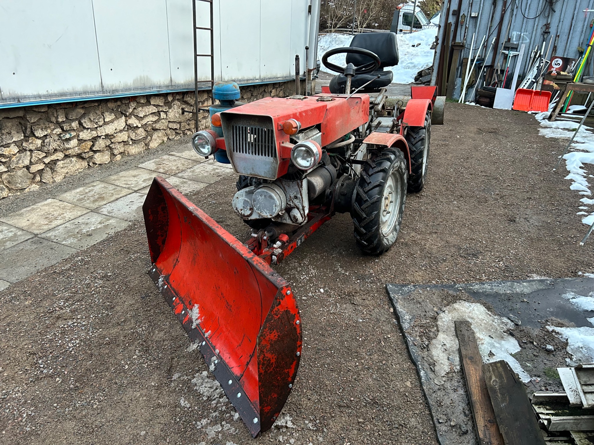 Czerwony, używany traktor z pługiem do śniegu stoi na żwirowym podłożu przed budynkiem z kamienną podstawą i blaszakiem ze sprzętem.