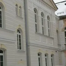 Odnowiona, jasna elewacja budynku z charakterystycznymi łukowymi oknami, widoczny fragment dachu pokrytego śniegiem i samochód również pod śniegiem.