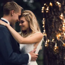 Para młoda w objęciach przed drzewem ozdobionym girlandą świetlną; panna młoda ma upięte włosy z wplecionym warkoczem i koronkową suknię z odkrytymi plecami.