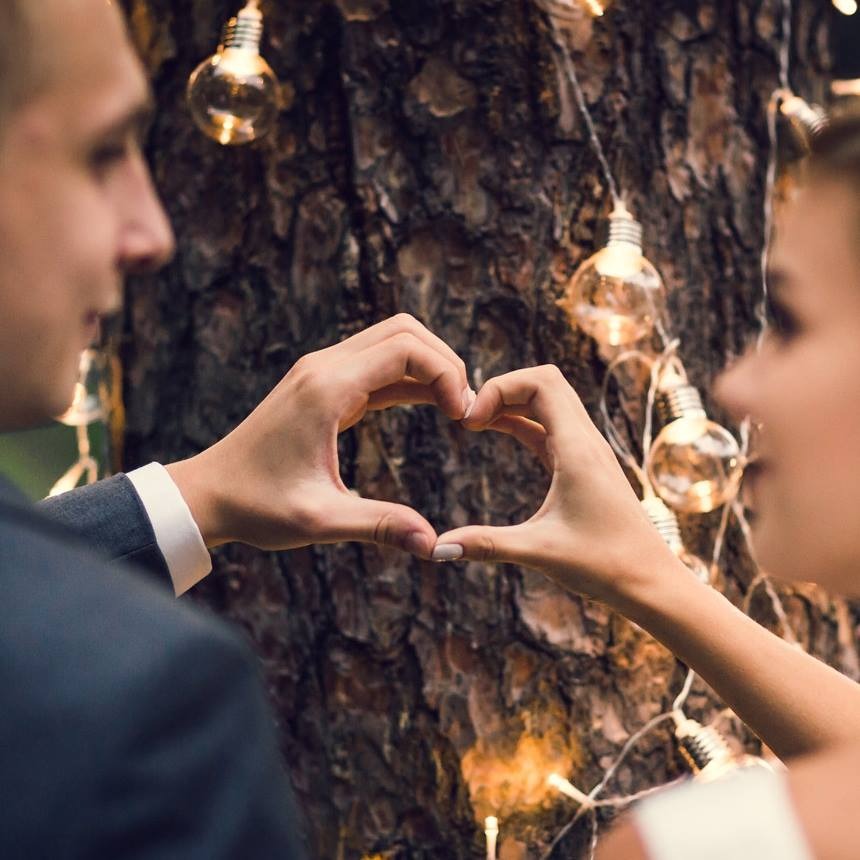 Para młoda tworzy serce dłońmi na tle kory drzewa ozdobionego girlandą z żarówek, widoczny profil twarzy panny młodej i fragment marynarki pana młodego.