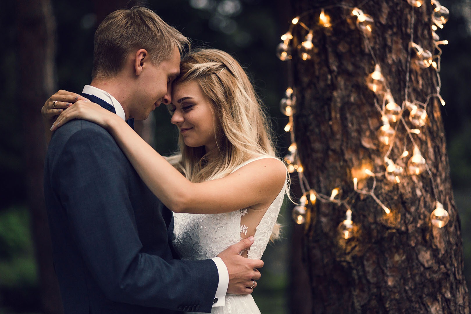 Para młoda w objęciach przed drzewem ozdobionym girlandą świetlną; panna młoda ma upięte włosy z wplecionym warkoczem i koronkową suknię z odkrytymi plecami.