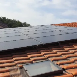 Panel słoneczny zamontowany na dachu z pomarańczowej dachówki, widoczny świetlik dachowy i dom w tle.