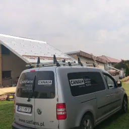 Srebrny samochód dostawczy z logo firmy Satfans i Canal+ na tle budynku z nowym, metalowym dachem i stertą desek. Na dachu samochodu zamontowany bagażnik z drabiną.
