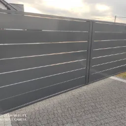 Nowoczesna, przesuwna brama wjazdowa w kolorze grafitowym z poziomymi panelami, zamontowana na posesji z kostką brukową, widoczny fragment domu w tle.