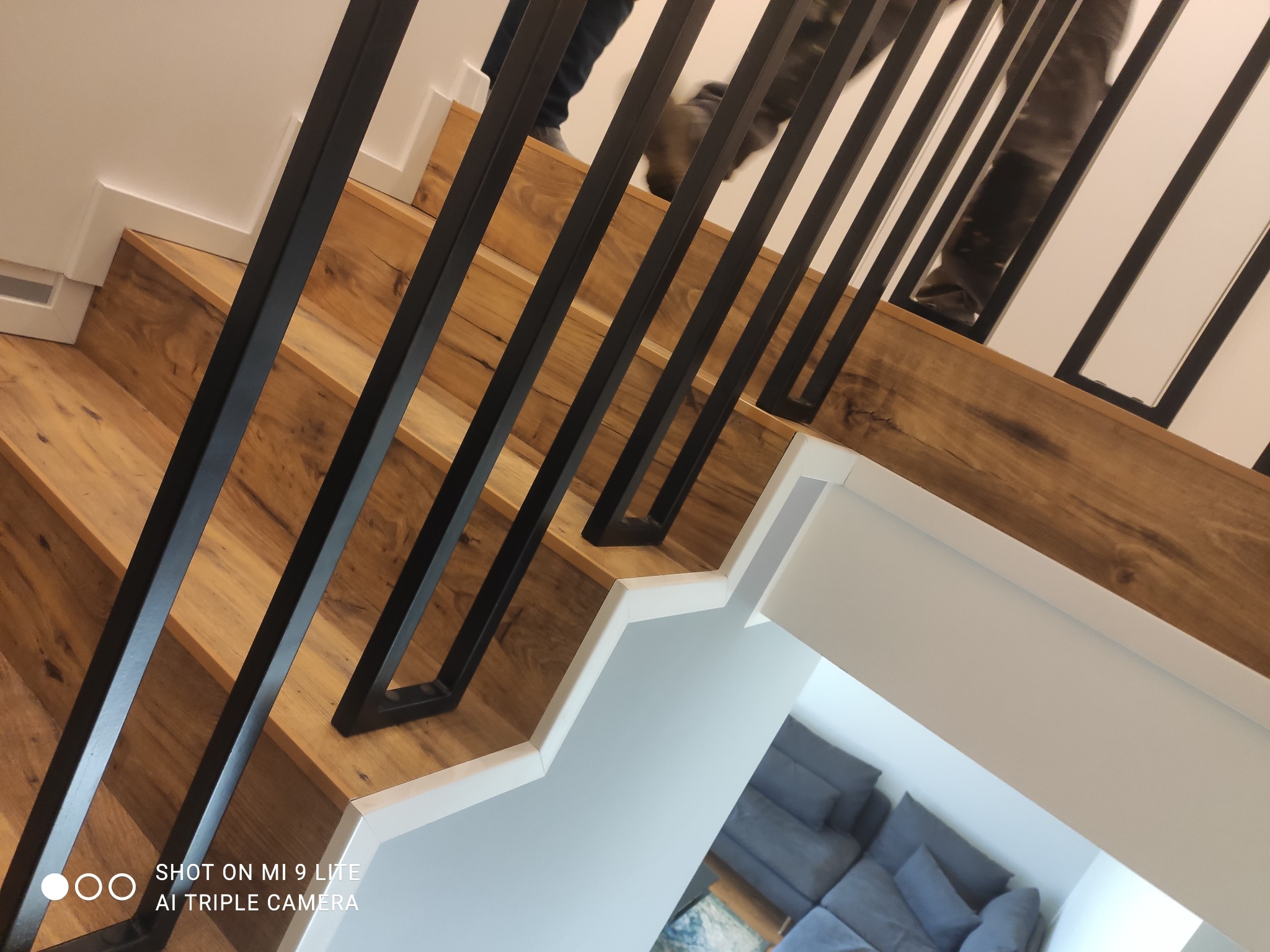 Schody drewniane z czarną, metalową balustradą, widok z góry z fragmentem osoby schodzącej, jasne wnętrze.
