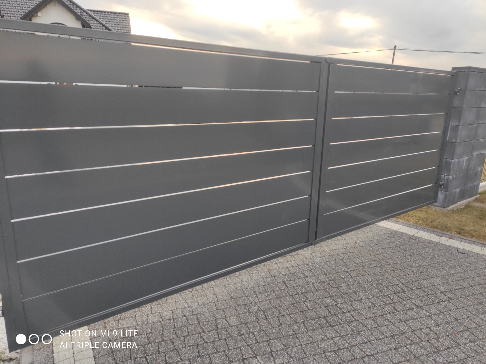 Nowoczesna, przesuwna brama wjazdowa w kolorze grafitowym z poziomymi panelami, zamontowana na posesji z kostką brukową, widoczny fragment domu w tle.