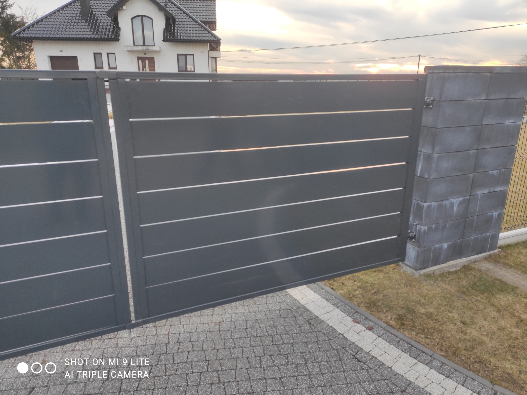 Nowoczesna, przesuwna brama wjazdowa z poziomymi panelami w kolorze grafitowym, zamontowana na posesji z kostką brukową i widocznym domem w tle, sfotografowana w pochmurny dzień.