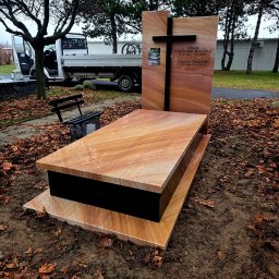 Kamieniarstwo Bieleccy - Nowoczesny nagrobek z piaskowca z czarnym krzyżem i tabliczką, otoczony jesiennymi liśćmi. W tle częściowo widoczny samochód dostawczy i ławka.