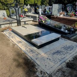 Kamieniarstwo Bieleccy - Granitowy nagrobek z czarną tablicą inskrypcyjną i krzyżem, otoczony kamienną kostką brukową. W tle inne nagrobki i zieleń cmentarza.