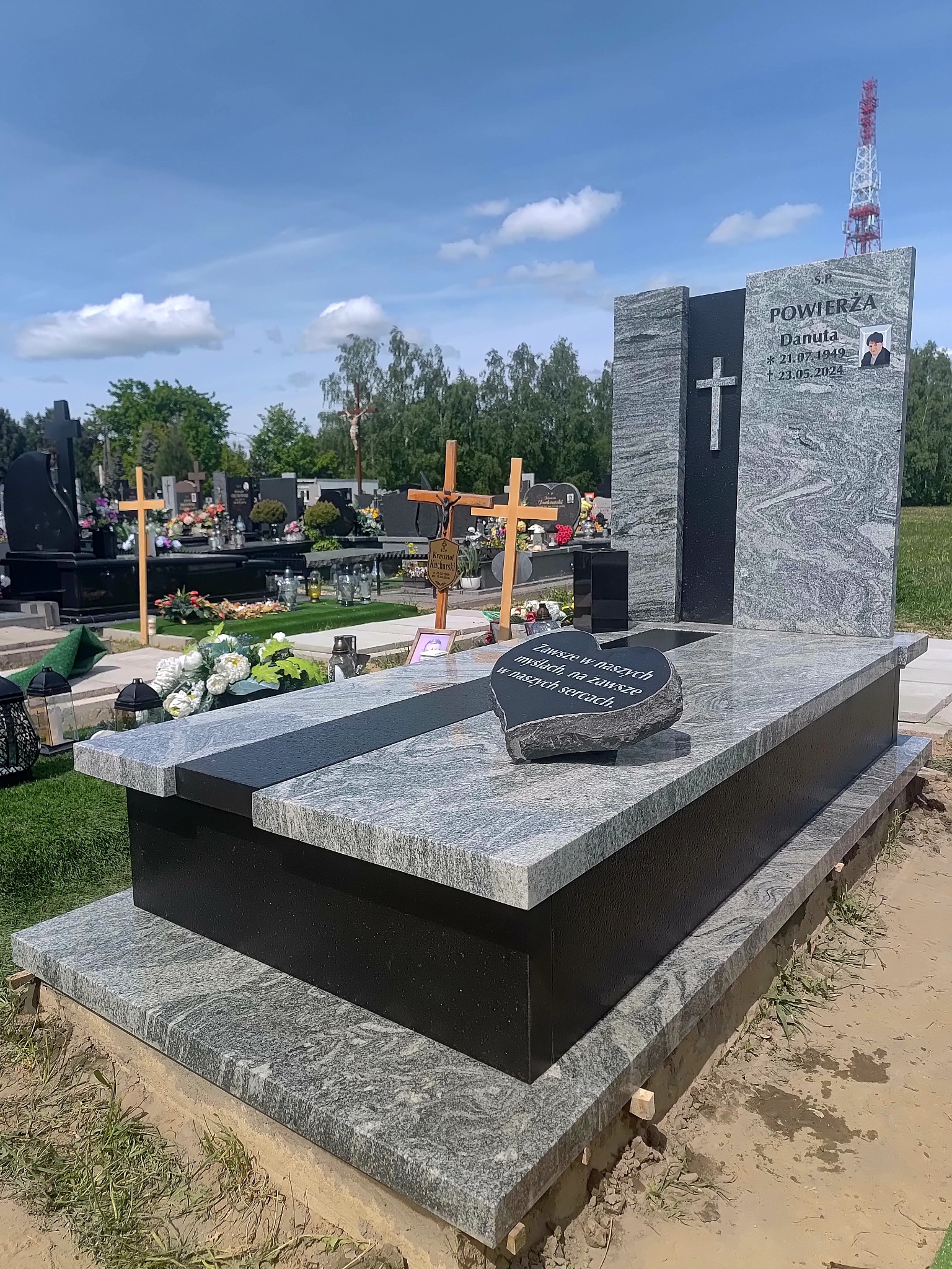 Granitowy nagrobek z czarnym akcentem i wyrytym napisem, otoczony innymi nagrobkami na tle błękitnego nieba i zieleni cmentarza. Widoczna wieża telekomunikacyjna w tle.