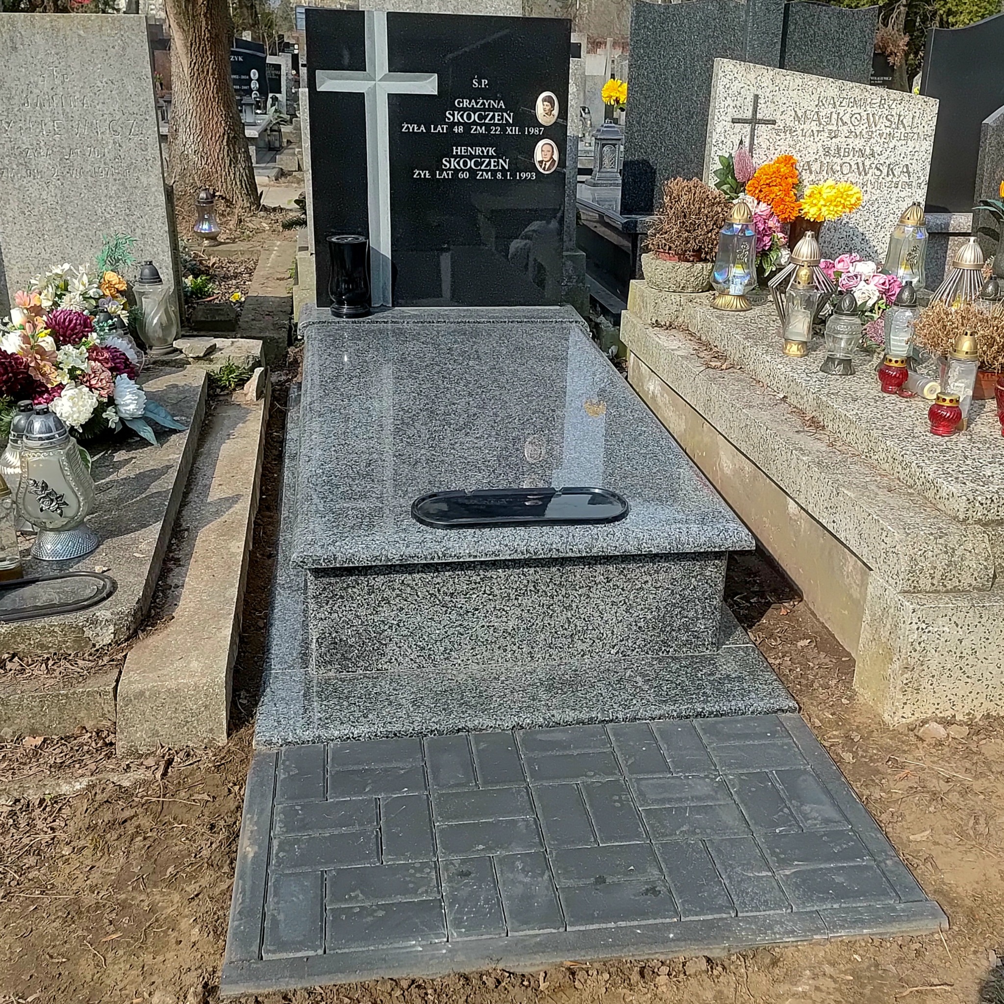 Granitowy nagrobek z czarną tablicą i zdjęciami zmarłych, otoczony kostką brukową. Widok na cmentarz z innymi nagrobkami w tle, w słoneczny dzień.