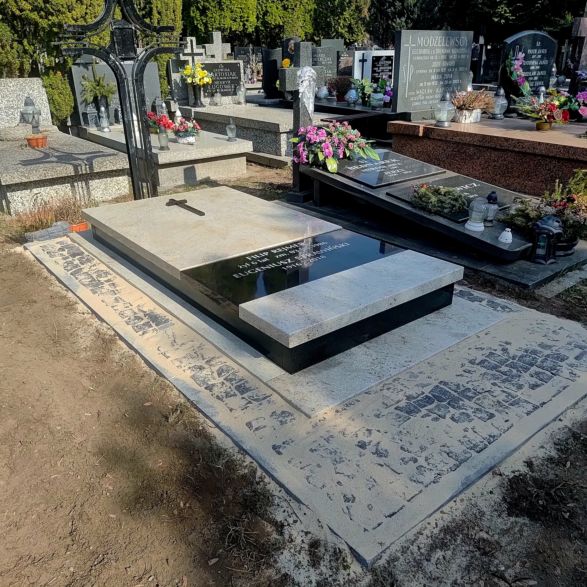 Granitowy nagrobek z czarną tablicą inskrypcyjną i krzyżem, otoczony kamienną kostką brukową. W tle inne nagrobki i zieleń cmentarza.