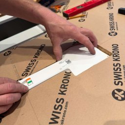 IE SOLUTIONS SPÓŁKA Z OGRANICZONĄ ODPOWIEDZIALNOŚCIĄ - Przygotowanie do montażu paneli podłogowych Swiss Krono: dłonie przytrzymują wzornik z próbką koloru z małą flagą, obok poziomica i ołówek.