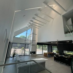 IE SOLUTIONS SPÓŁKA Z OGRANICZONĄ ODPOWIEDZIALNOŚCIĄ - Wysoki salon z oknami trapezowymi, oświetleniem LED w suficie, minimalistycznym żyrandolem i szarą sofą modułową, widok z antresoli.