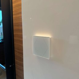 IE SOLUTIONS SPÓŁKA Z OGRANICZONĄ ODPOWIEDZIALNOŚCIĄ - Biały dotykowy panel sterowania oświetleniem z podświetleniem LED, zamontowany na gładkiej, białej ścianie, widoczny fragment drewnianej zabudowy.