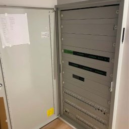 IE SOLUTIONS SPÓŁKA Z OGRANICZONĄ ODPOWIEDZIALNOŚCIĄ - Otwarta szafa elektryczna z listwami zaciskowymi oznaczonymi literami i cyframi, widoczna dokumentacja techniczną na wewnętrznej stronie drzwi.