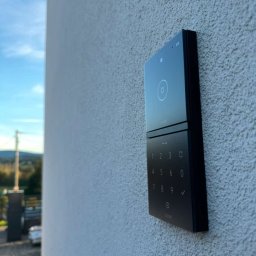 IE SOLUTIONS SPÓŁKA Z OGRANICZONĄ ODPOWIEDZIALNOŚCIĄ - Czarny, nowoczesny wideodomofon z klawiaturą numeryczną zamontowany na elewacji budynku o chropowatej strukturze, widoczna kamera monitoringu w górnym rogu i fragment krajobrazu w tle.