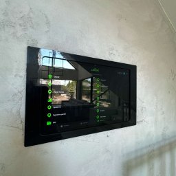 IE SOLUTIONS SPÓŁKA Z OGRANICZONĄ ODPOWIEDZIALNOŚCIĄ - Czarny panel sterowania Loxone zamontowany na szarej ścianie o nierównej fakturze, wyświetlający menu z ikonami i nazwami pomieszczeń, z widocznym odbiciem okna i fragmentu wnętrza w ekranie.