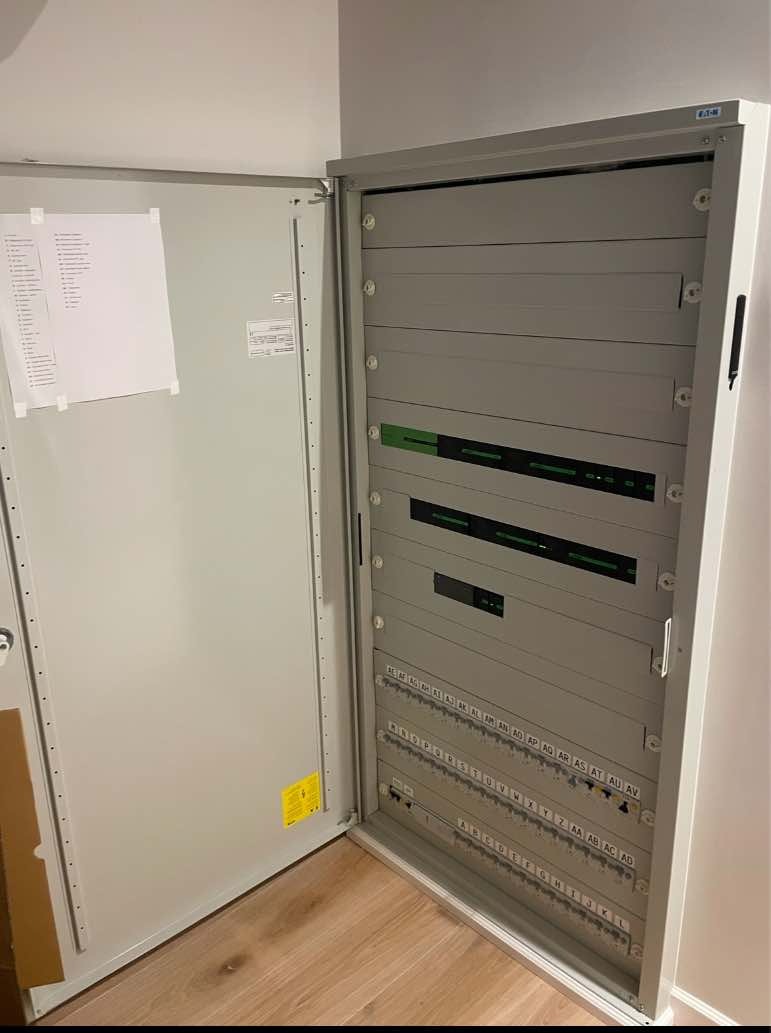 Otwarta szafa elektryczna z listwami zaciskowymi oznaczonymi literami i cyframi, widoczna dokumentacja techniczną na wewnętrznej stronie drzwi.