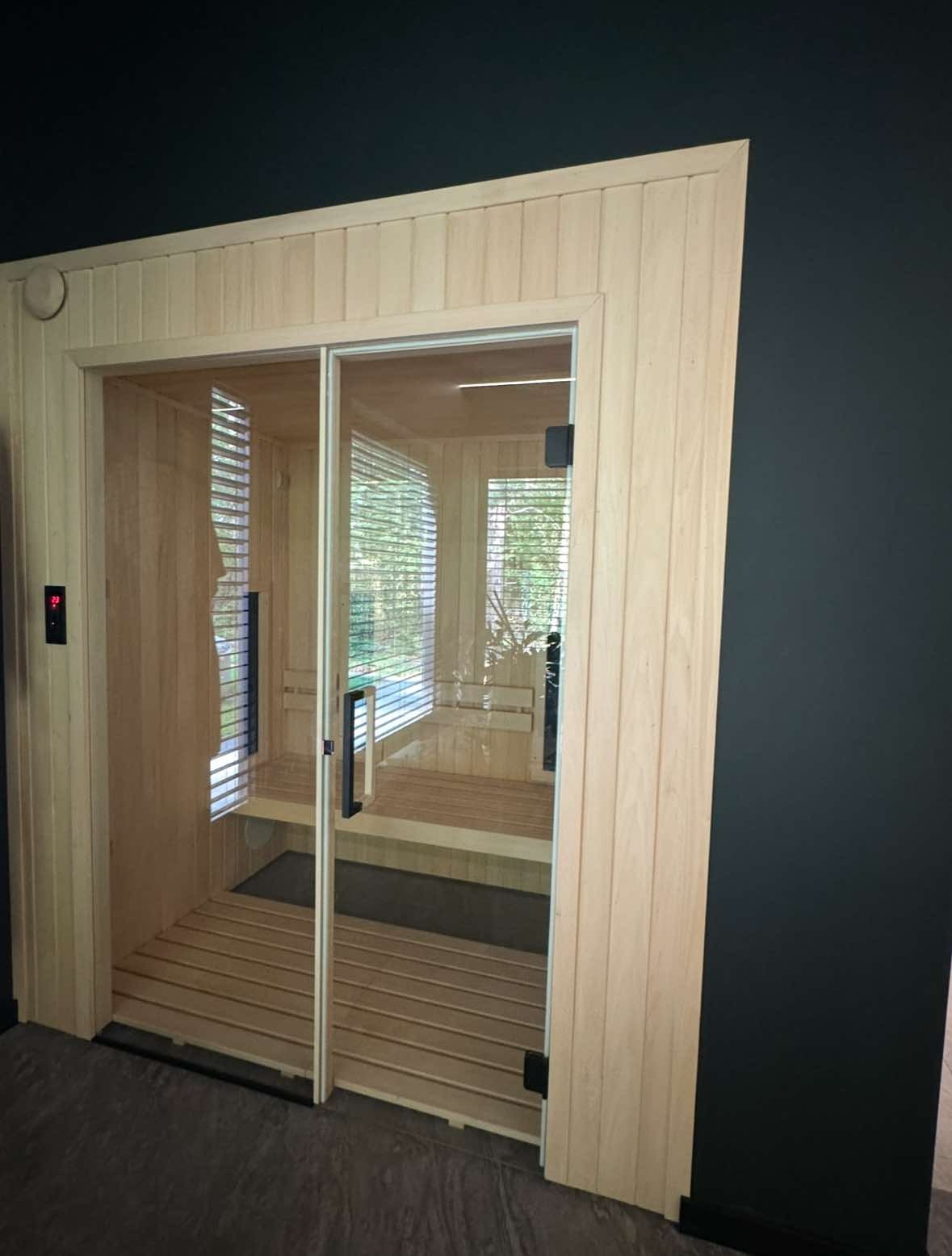 Gotowa sauna w pomieszczeniu, widok z zewnątrz przez szklane drzwi przesuwne, drewniane wykończenie i ławki, elektroniczny panel sterowania temperatury po lewej stronie.